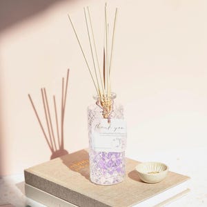Puede incluir: Un difusor de aromaterapia de vidrio transparente con un patrón de diamantes, lleno de líquido púrpura y cañas. Una etiqueta dice "gracias". El difusor está sobre una pila de libros, junto a un pequeño cuenco blanquecino.