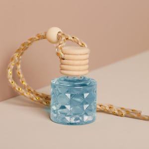 Puede incluir: Pequeña botella de vidrio azul claro con un patrón de diamantes, tapa de madera y cuerda dorada y beige. La botella está diseñada para colgar y tiene una cuenta de madera beige claro en la parte superior de la cuerda.