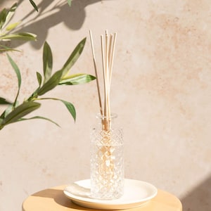 May include: A decorative glass diffuser bottle with a diamond pattern, filled with reeds, sits on a small white plate. The bottle is on a light wood stool. Olive branches are in the background.