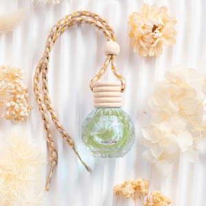 May include: A spherical glass car air freshener with a pale green liquid, wooden cap, and a braided rope with gold detailing. The background is white fabric with dried flowers. The freshener is designed to hang.