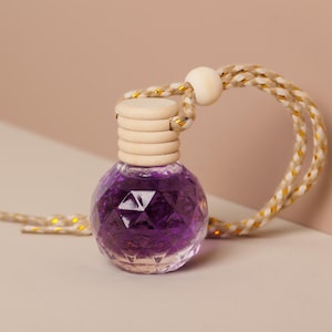 Puede incluir: Pequeña botella de vidrio facetado llena de líquido morado oscuro. Tiene una tapa de madera y está suspendida por una cuerda con detalles dorados. Diseño simple y elegante, adecuado para aromaterapia o fragancia.
