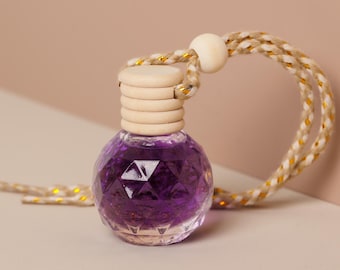 May include: A small, faceted glass bottle filled with a deep purple liquid. The bottle has a wooden cap and is suspended by a rope with gold accents. The design is simple and elegant, suitable for aromatherapy or fragrance.