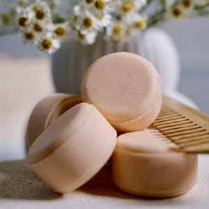 May include: A close-up of several light peach-colored, round solid shampoo bars. A wooden comb rests on the side. White flowers in a white vase are in the background, suggesting a spa or self-care theme.