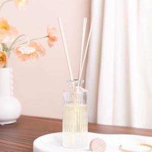 Puede incluir: Un frasco difusor de vidrio transparente con estrías verticales, lleno de un líquido amarillo claro y varillas de madera. El frasco está sobre una bandeja de mármol blanco, junto a una tapa de madera y una pulsera dorada. Flores color melocotón en el fondo.