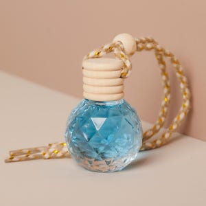 Puede incluir: Una pequeña botella redonda de vidrio azul con un patrón geométrico. La botella tiene una tapa de madera y está suspendida por una cuerda con detalles dorados. El fondo es de un color suave y neutro.