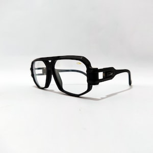 Puede incluir: Gafas con montura negra con un diseño angular distintivo. Las gafas tienen una montura gruesa y audaz y lentes transparentes. La estética general es moderna y elegante, adecuada para el uso diario. Las gafas están sobre un fondo blanco liso.
