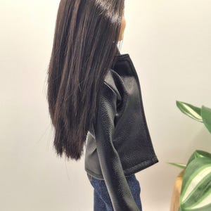 May include: A doll wearing a black faux leather jacket and blue denim jeans. The doll has long, dark brown hair. The jacket has a classic biker style with a notched collar and a slightly textured appearance. The jeans are a medium wash.