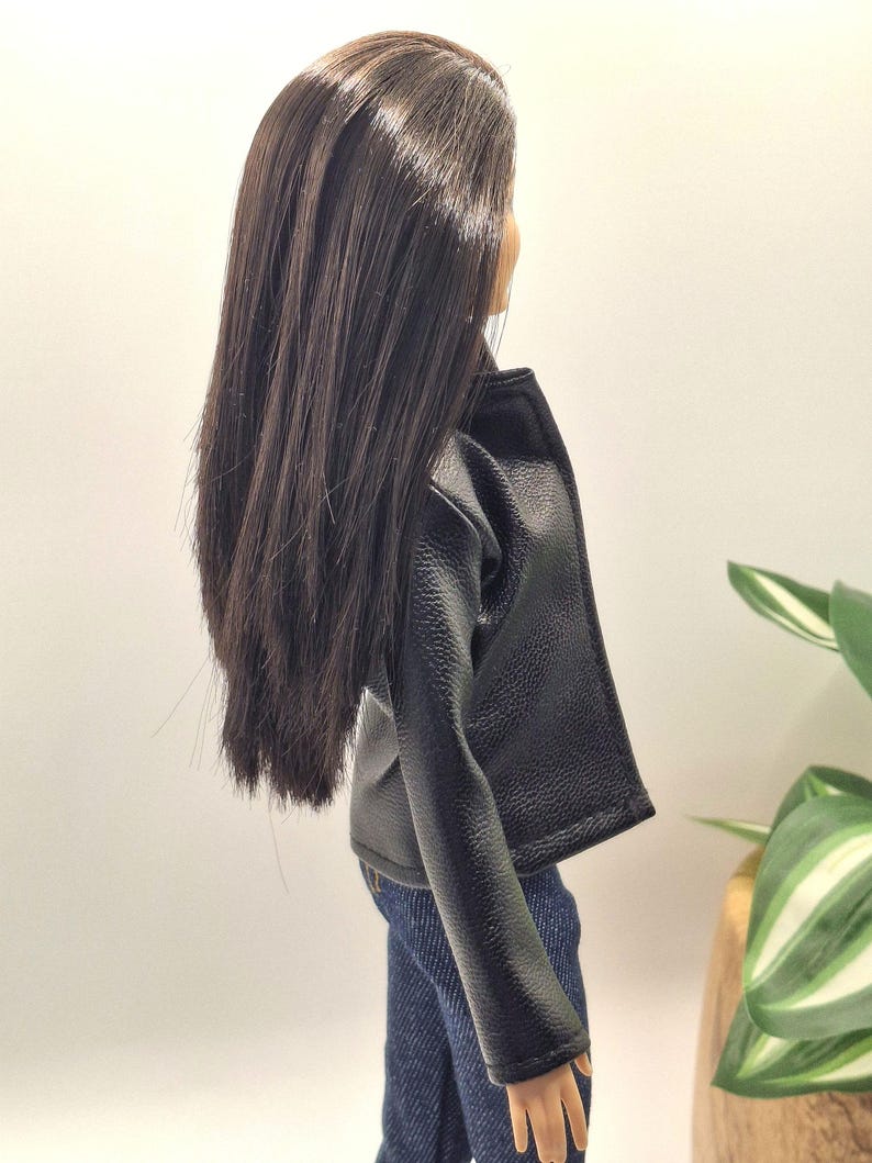 May include: A doll wearing a black faux leather jacket and blue denim jeans. The doll has long, dark brown hair. The jacket has a classic biker style with a notched collar and a slightly textured appearance. The jeans are a medium wash.