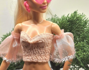 May include: A doll wearing pink heart-shaped sunglasses and matching hoop earrings. The doll is also wearing a light pink off-the-shoulder top with sheer sleeves and light blue knit pants. The doll has blonde hair.