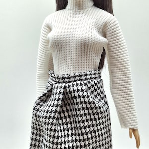 May include: A doll wearing a white turtleneck sweater and a black and white houndstooth skirt. The skirt has a wrap-around design. The doll has long, dark hair and is wearing pearl earrings.