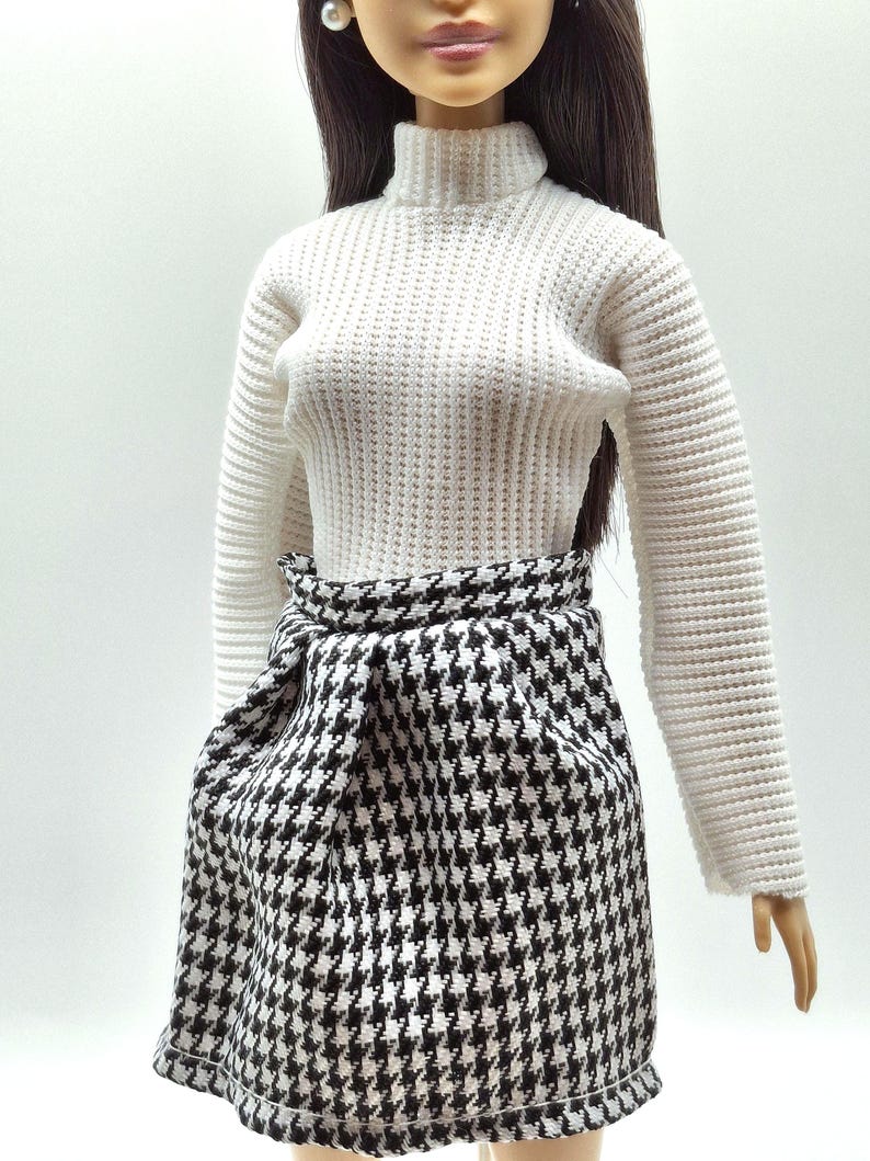 May include: A doll wearing a white turtleneck sweater and a black and white houndstooth skirt. The skirt has a wrap-around design. The doll has long, dark hair and is wearing pearl earrings.