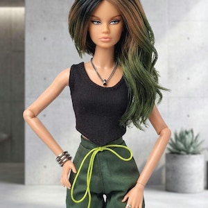 May include: A fashion doll with long, wavy hair transitioning from black to green. The doll wears a black sleeveless top, dark green cargo pants with a bright neon yellow drawstring waist, a silver chain necklace, and a silver-toned multi-band bracelet.