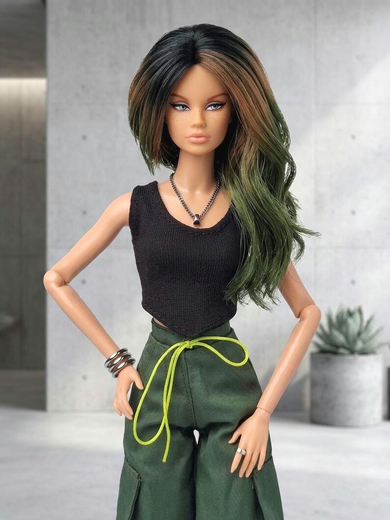 May include: A fashion doll with long, wavy hair transitioning from black to green. The doll wears a black sleeveless top, dark green cargo pants with a bright neon yellow drawstring waist, a silver chain necklace, and a silver-toned multi-band bracelet.