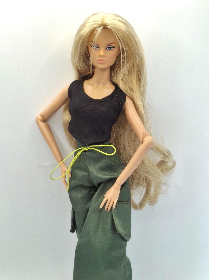 May include: A fashion doll with long blonde hair, wearing a black sleeveless top and olive green cargo pants with large side pockets. The pants feature a bright neon yellow drawstring at the waist. The doll has articulated limbs.