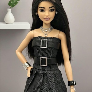 K&ouml;nnte beinhalten: Eine Puppe mit langem dunklem Haar tr&auml;gt ein schwarzes tr&auml;gerloses Kleid mit zwei horizontalen schwarzen G&uuml;rteln mit silbernen Schnallen. Die Puppe ist mit einer mehrlagigen silbernen Halskette, silbernen Ohrsteckern, einem Armreif und einem Armband versehen.
