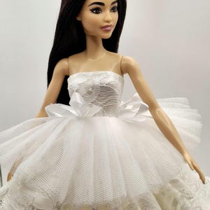 May include: A white strapless dress with a lace bodice and a tulle skirt. The dress has a bow at the waist and is worn by a doll with long black hair.