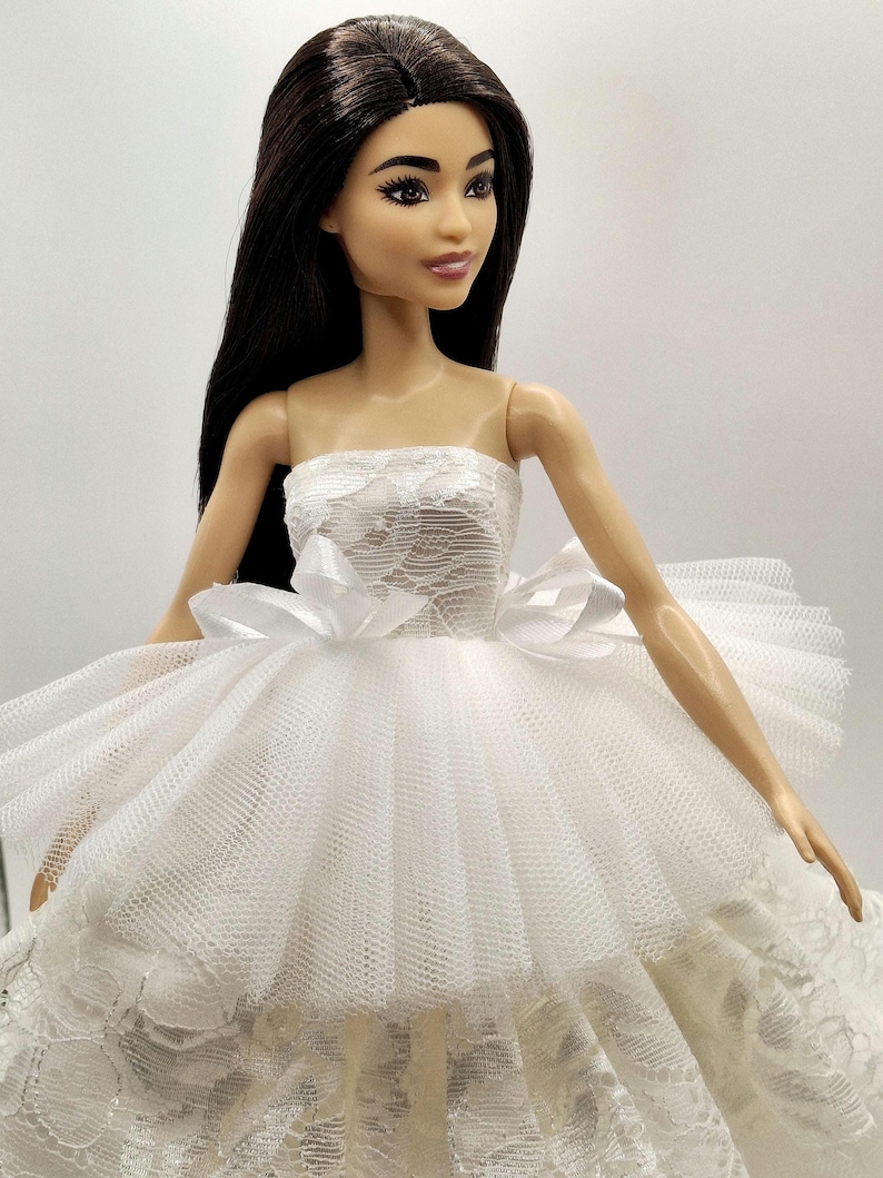May include: A white strapless dress with a lace bodice and a tulle skirt. The dress has a bow at the waist and is worn by a doll with long black hair.
