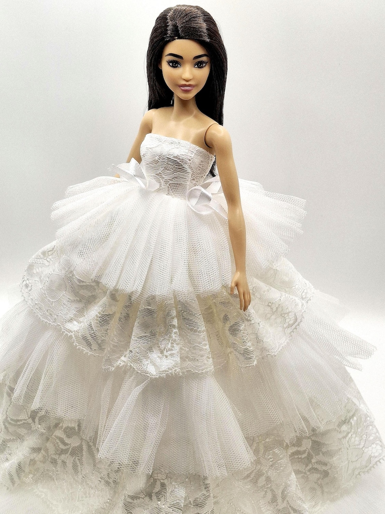 May include: A white strapless doll dress with a lace bodice and tulle skirt. The dress has multiple layers of tulle and lace, creating a voluminous and elegant look.
