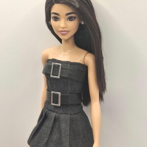 K&ouml;nnte beinhalten: Eine Puppe mit langem, glattem, dunkelbraunem Haar und braunen Augen, bekleidet mit einem tr&auml;gerlosen, schwarzen, strukturierten Minikleid. Das Kleid hat zwei horizontale schwarze Kunstledergurte am Oberteil, jeweils mit einer silbernen Schnalle.