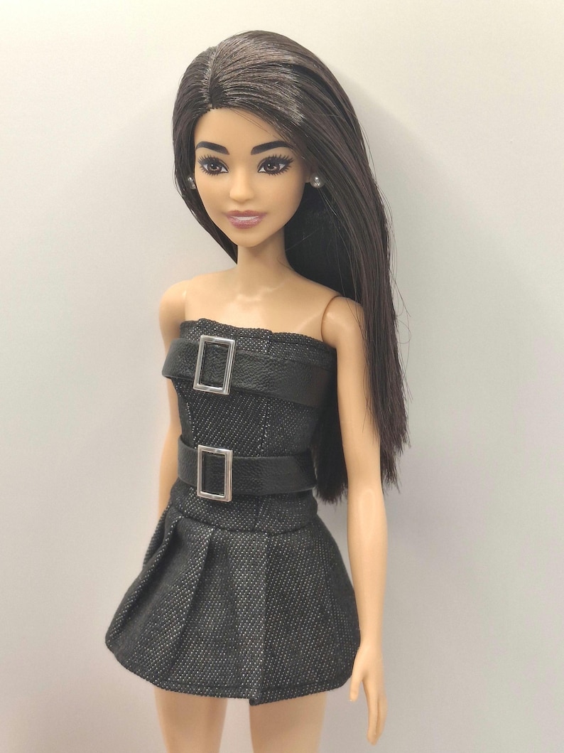 K&ouml;nnte beinhalten: Eine Puppe mit langem, glattem, dunkelbraunem Haar und braunen Augen, bekleidet mit einem tr&auml;gerlosen, schwarzen, strukturierten Minikleid. Das Kleid hat zwei horizontale schwarze Kunstledergurte am Oberteil, jeweils mit einer silbernen Schnalle.