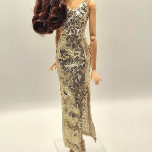 May include: A doll wearing a gold sequined evening gown with a V-neck and a thigh-high slit. The doll has long, auburn hair and is wearing matching gold heels. The dress is embellished with gold sequins.