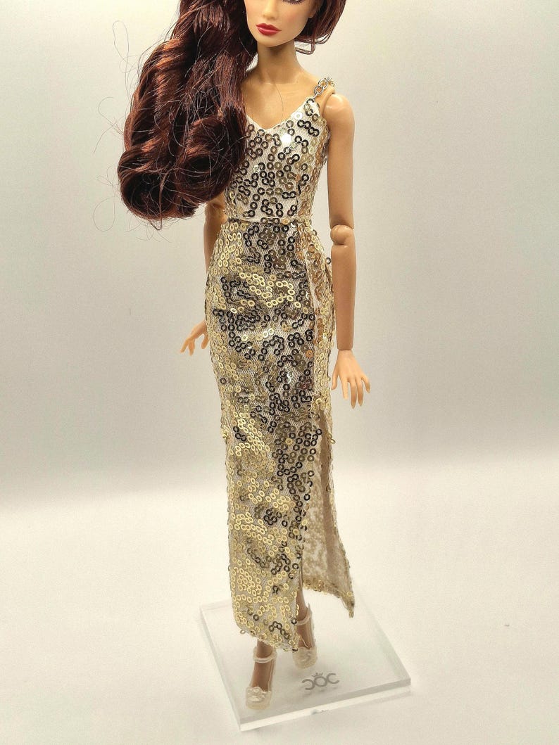 May include: A doll wearing a gold sequined evening gown with a V-neck and a thigh-high slit. The doll has long, auburn hair and is wearing matching gold heels. The dress is embellished with gold sequins.