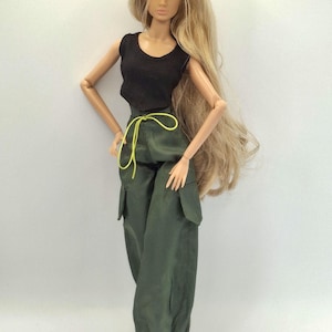 May include: A fashion doll with long blonde hair wearing a black sleeveless top and olive green cargo pants featuring side pockets, a cinched waist with a bright yellow drawstring, and cuffed ankles, paired with black shoes.