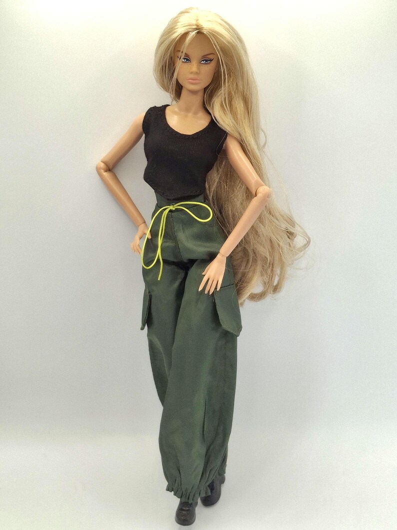 May include: A fashion doll with long blonde hair wearing a black sleeveless top and olive green cargo pants featuring side pockets, a cinched waist with a bright yellow drawstring, and cuffed ankles, paired with black shoes.