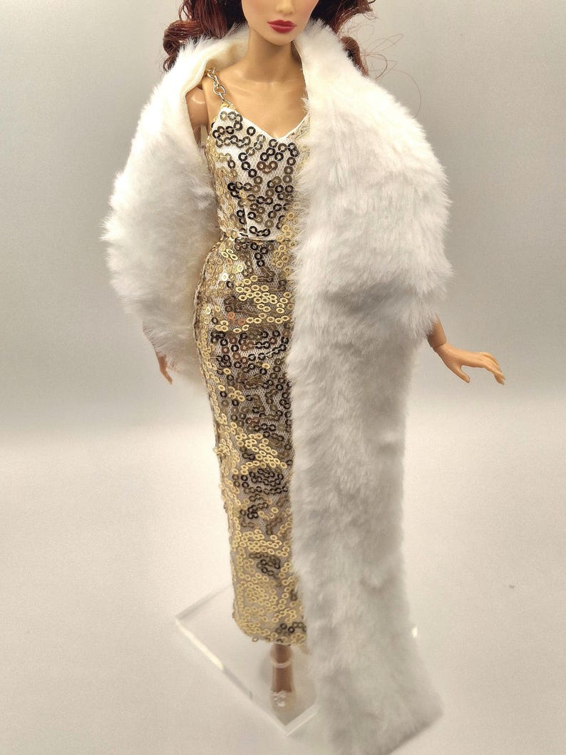 May include: A doll wearing a gold sequined dress and a white faux fur stole. The dress has a V-neck and spaghetti straps. The stole is draped over the shoulders. The doll has red hair and is standing on a clear stand.