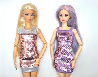 May include: Two fashion dolls are displayed, each wearing a sequined mini-dress. One doll has blonde hair and a pink dress, while the other has purple hair and a purple dress. Both dresses have a silver trim.
