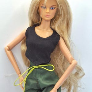 May include: A fashion doll with long blonde hair, blue eyes, and freckles. The doll wears a sleeveless black top and olive green trousers featuring a bright yellow drawstring waist tie. The doll has articulated arms and a neutral facial expression.