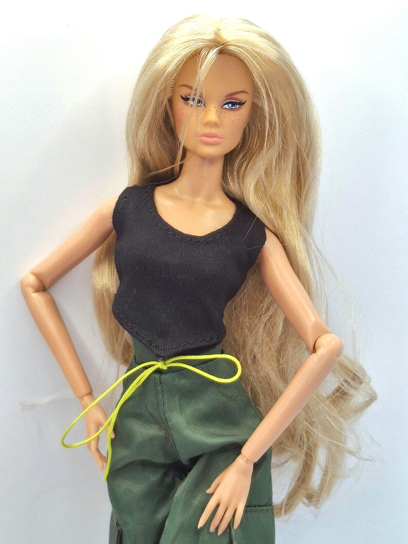 May include: A fashion doll with long blonde hair, blue eyes, and freckles. The doll wears a sleeveless black top and olive green trousers featuring a bright yellow drawstring waist tie. The doll has articulated arms and a neutral facial expression.