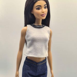 May include: A fashion doll with long black hair, wearing a white sleeveless top with a black trim and dark blue denim pants. The doll has a neutral expression and is posed against a plain background.