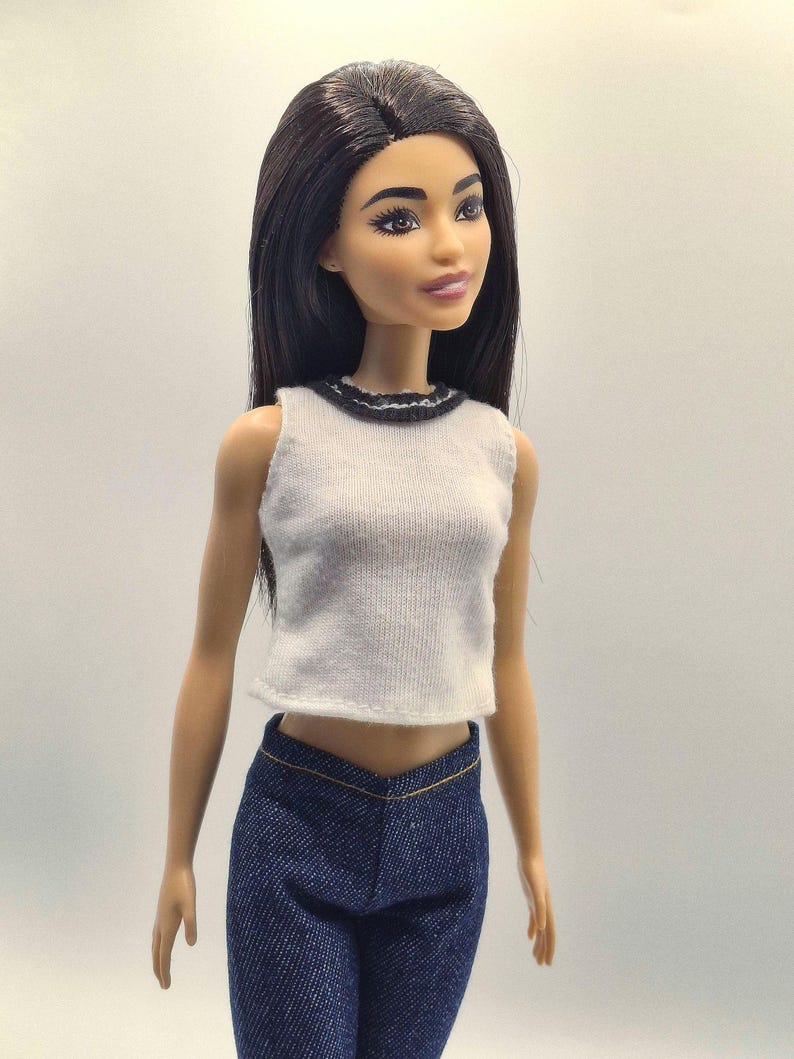 May include: A fashion doll with long black hair, wearing a white sleeveless top with a black trim and dark blue denim pants. The doll has a neutral expression and is posed against a plain background.