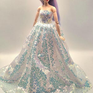 K&ouml;nnte beinhalten: Puppe mit langem, lavendelfarbenem Haar und einem tr&auml;gerlosen Kleid. Das wei&szlig;e Kleid ist mit schillernden Pailletten bedeckt, die blau und gr&uuml;n schimmern. Die Puppe hat ein zartes Gesicht und steht vor einem schlichten Hintergrund.
