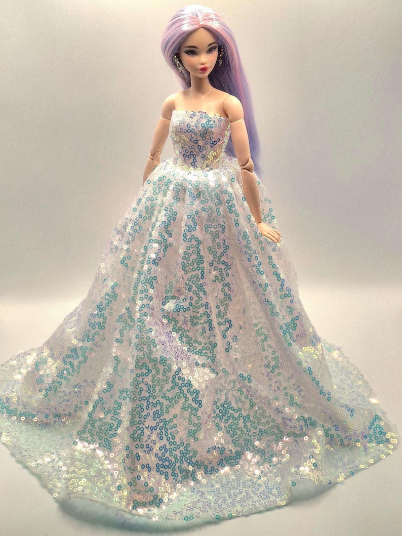 K&ouml;nnte beinhalten: Puppe mit langem, lavendelfarbenem Haar und einem tr&auml;gerlosen Kleid. Das wei&szlig;e Kleid ist mit schillernden Pailletten bedeckt, die blau und gr&uuml;n schimmern. Die Puppe hat ein zartes Gesicht und steht vor einem schlichten Hintergrund.