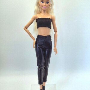 May include: A doll with blonde hair styled in a bun, wearing a black tube top, black pants, and black ankle boots. The doll is standing on a clear plastic base.