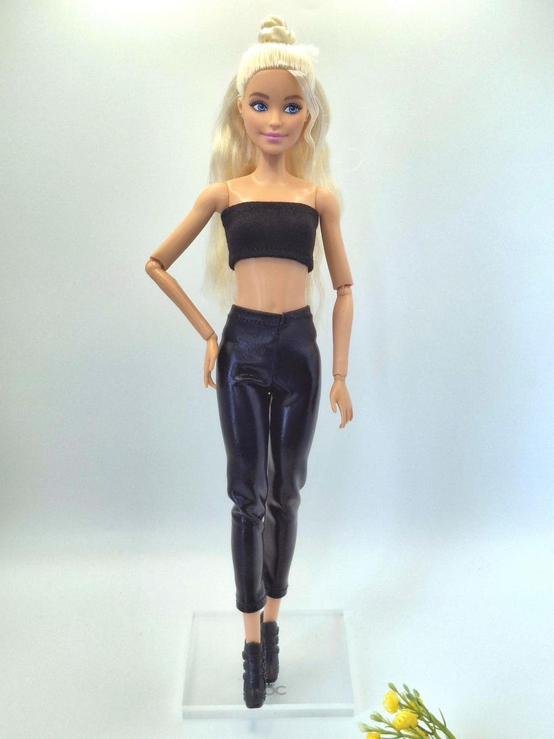 May include: A doll with blonde hair styled in a bun, wearing a black tube top, black pants, and black ankle boots. The doll is standing on a clear plastic base.