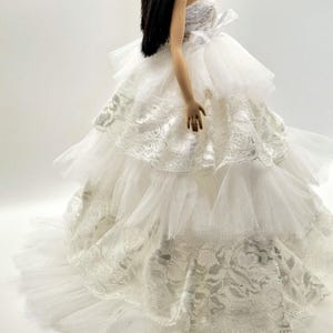 May include: A white tulle and lace wedding dress with multiple layers and a fitted bodice. The dress has a long train and is perfect for a doll.