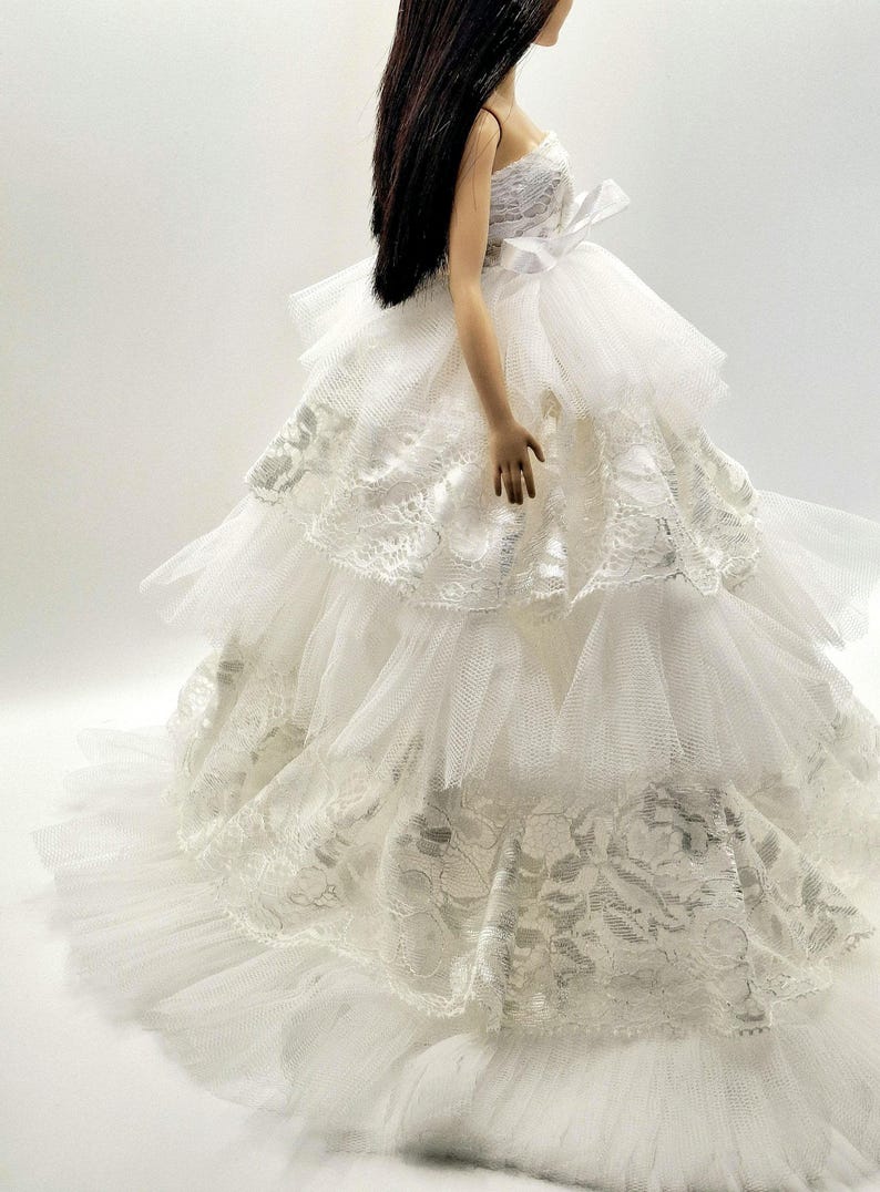 May include: A white tulle and lace wedding dress with multiple layers and a fitted bodice. The dress has a long train and is perfect for a doll.
