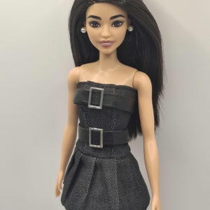 K&ouml;nnte beinhalten: Eine Puppe mit langem, glattem, dunkelbraunem Haar und heller Haut, die ein tr&auml;gerloses Kleid in schwarzer Denim-Optik tr&auml;gt. Das Kleid hat zwei horizontale schwarze Riemen an Brust und Taille, jeweils mit einer silbernen Schnalle.