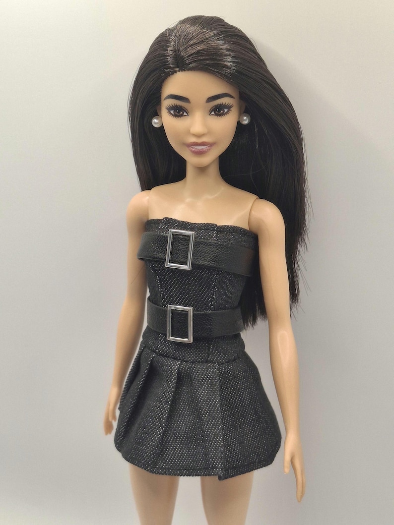 K&ouml;nnte beinhalten: Eine Puppe mit langem, glattem, dunkelbraunem Haar und heller Haut, die ein tr&auml;gerloses Kleid in schwarzer Denim-Optik tr&auml;gt. Das Kleid hat zwei horizontale schwarze Riemen an Brust und Taille, jeweils mit einer silbernen Schnalle.