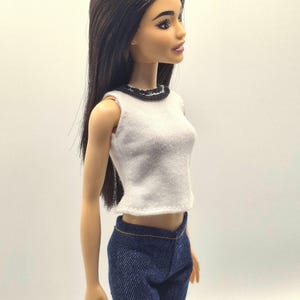 May include: A doll with long, dark hair wearing a white sleeveless top with a black trim and blue denim pants. The doll is posed in a side profile, showcasing the outfit's details.