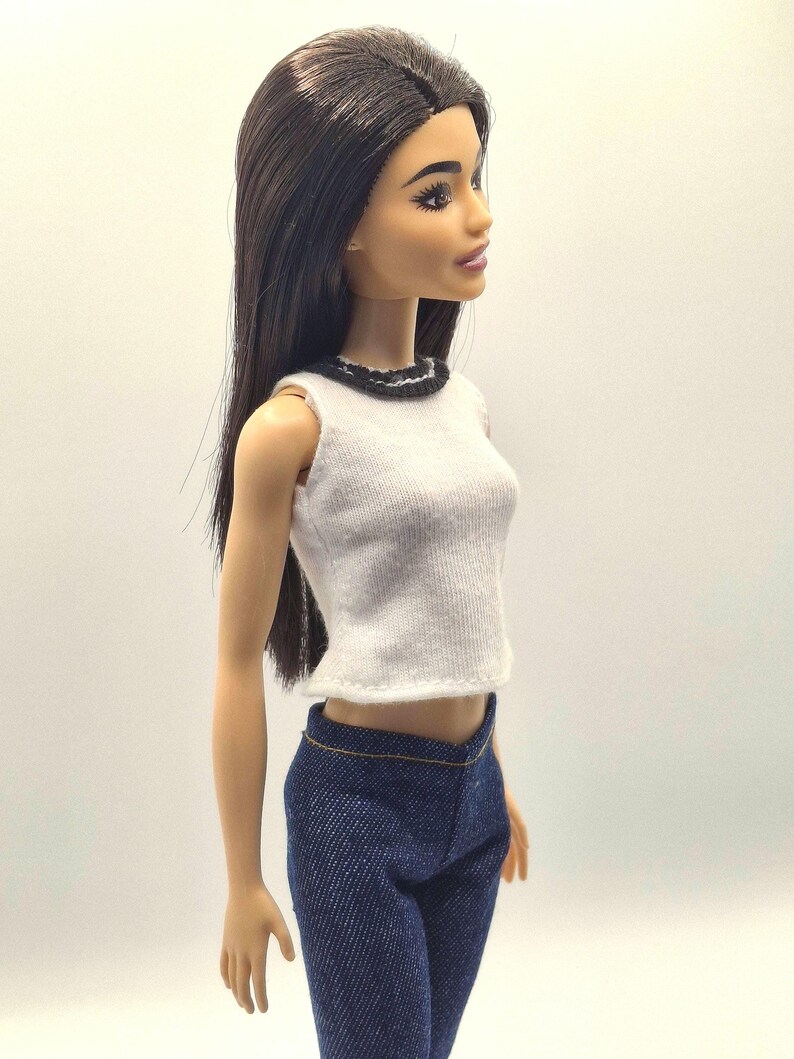 May include: A doll with long, dark hair wearing a white sleeveless top with a black trim and blue denim pants. The doll is posed in a side profile, showcasing the outfit's details.