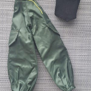 May include: Olive green cargo pants featuring side pockets, a high waist with a yellow drawstring, and elasticized cuffs, paired with a sleeveless black top. These doll-sized garments are crafted from a smooth, synthetic fabric.
