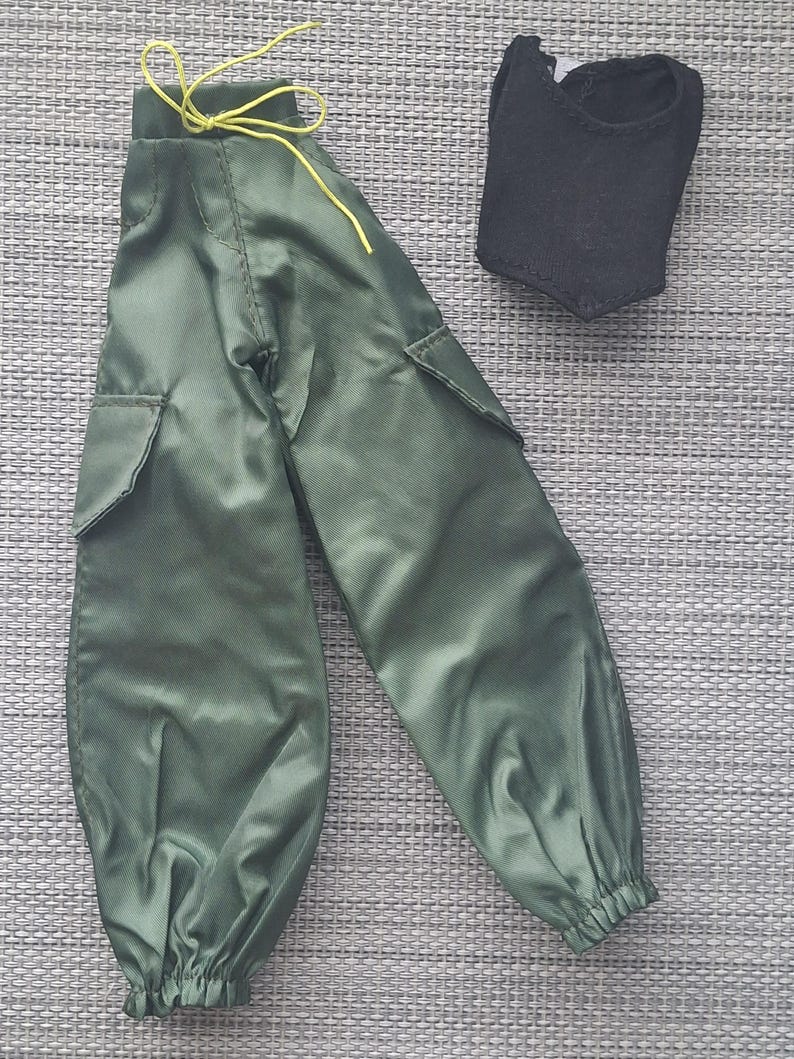 May include: Olive green cargo pants featuring side pockets, a high waist with a yellow drawstring, and elasticized cuffs, paired with a sleeveless black top. These doll-sized garments are crafted from a smooth, synthetic fabric.