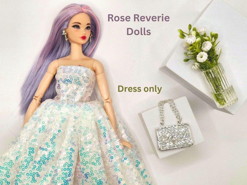 K&ouml;nnte beinhalten: Eine Puppe in einem tr&auml;gerlosen wei&szlig;en Kleid mit blauen und t&uuml;rkisfarbenen Pailletten. Die Puppe hat langes lavendelfarbenes Haar und tr&auml;gt Ohrringe. Eine kleine, glitzernde Handtasche und eine Vase mit wei&szlig;en Blumen sind ebenfalls im Bild. Der Text lautet "Rose Reverie Dolls" und "Dress only."