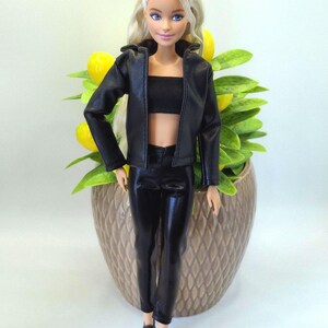 May include: A doll with blonde hair and blue eyes, wearing a black leather jacket, crop top, and pants. The doll is standing in front of a decorative pot with yellow lemons and green leaves.
