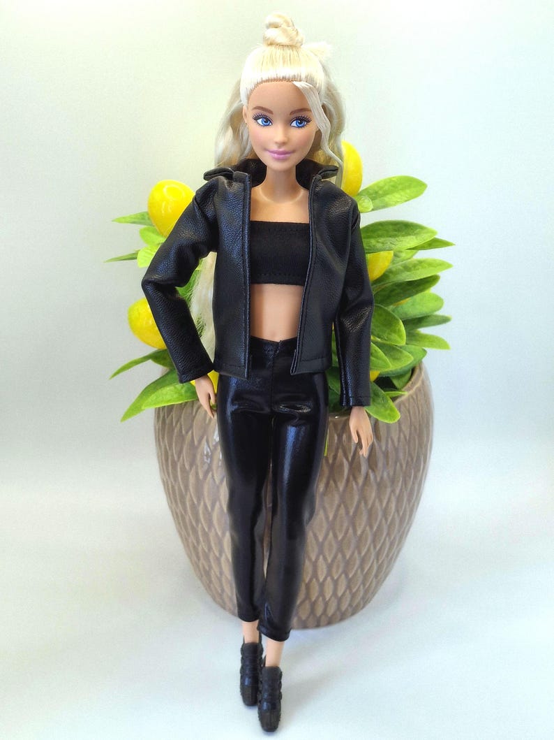 May include: A doll with blonde hair and blue eyes, wearing a black leather jacket, crop top, and pants. The doll is standing in front of a decorative pot with yellow lemons and green leaves.