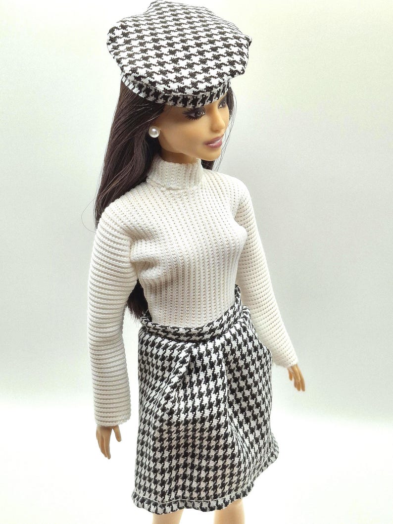 May include: A doll wearing a white turtleneck sweater, a black and white houndstooth skirt, and a matching houndstooth cap. The doll has long brown hair and a pearl earring. The outfit is a classic, stylish ensemble.